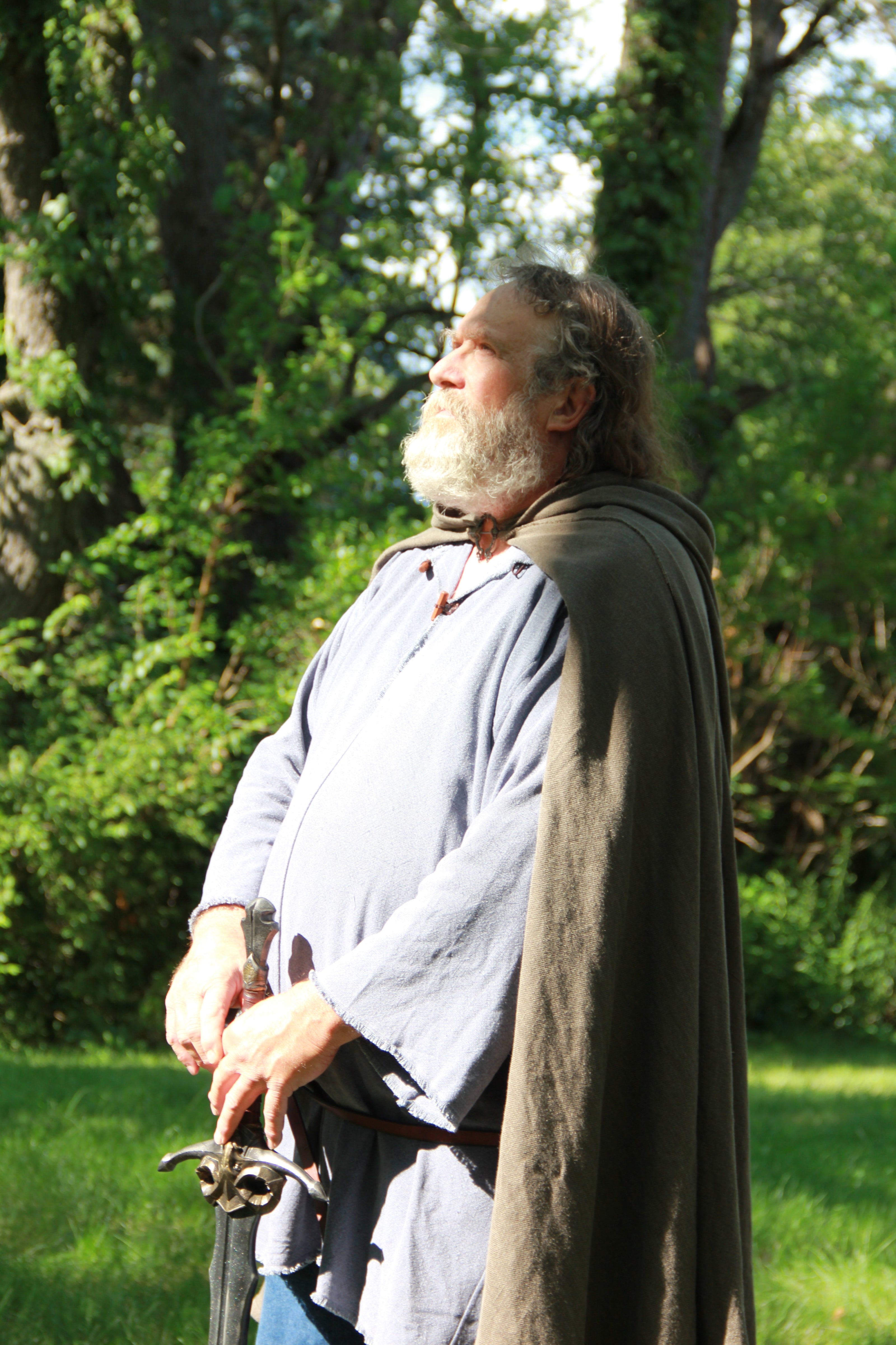 A bearded man dressed as Aragorn stands in the woods gazing into the distance as sunlight hits him. He is wearing a muted green cloak, a linen replica of the fellowship cloaks from lord of the rings. He also wears a muted blue tunic and holds a sword in front of him, resting the tip on the ground.