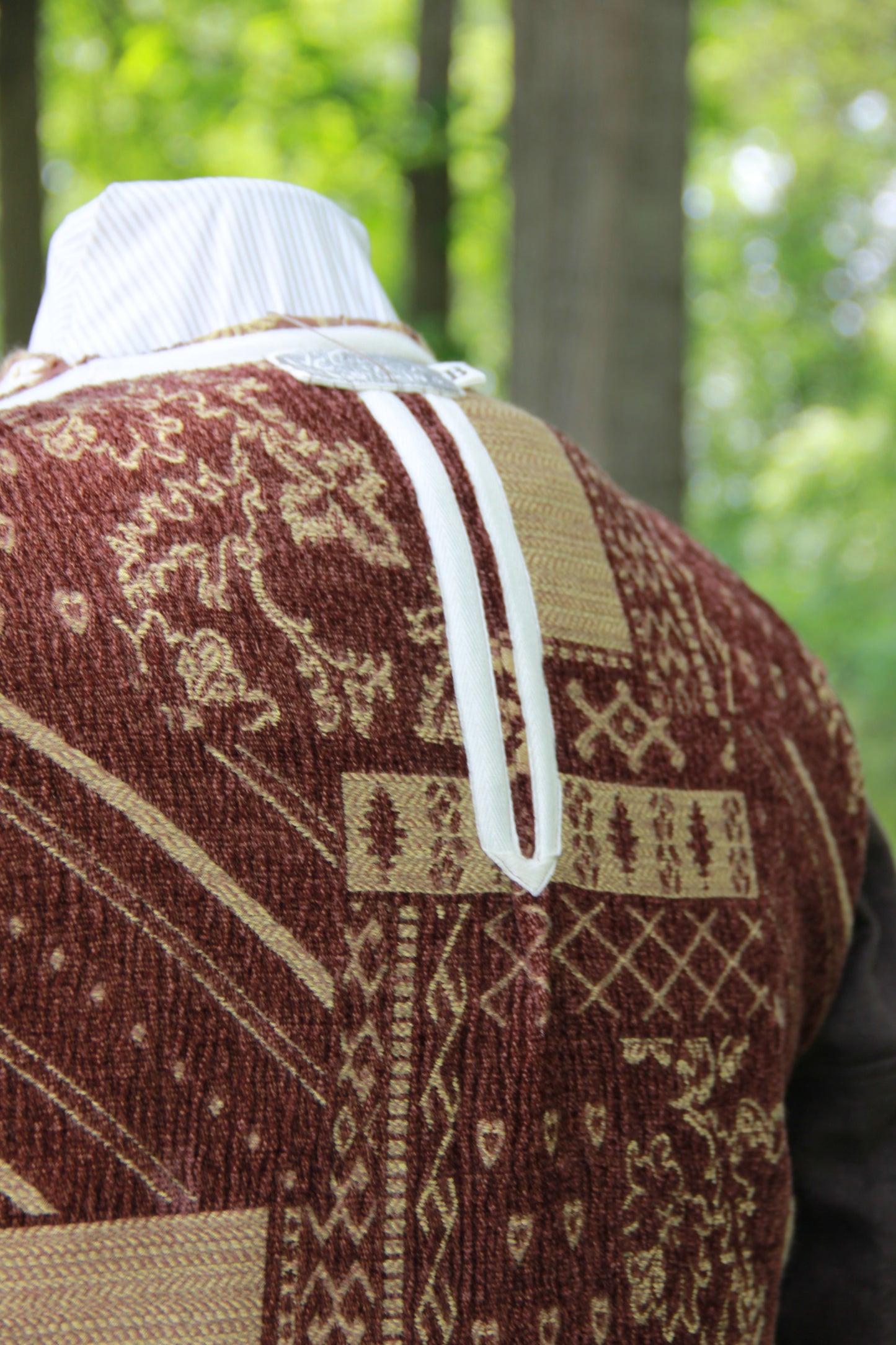 A close up of a hand made brown patterned chenille medieval jerkin at the back. It is inside out to display the ivory twill ribbon running along all of the seams, for decor and function. It is displayed on a masculine dress form in a woodland scene.