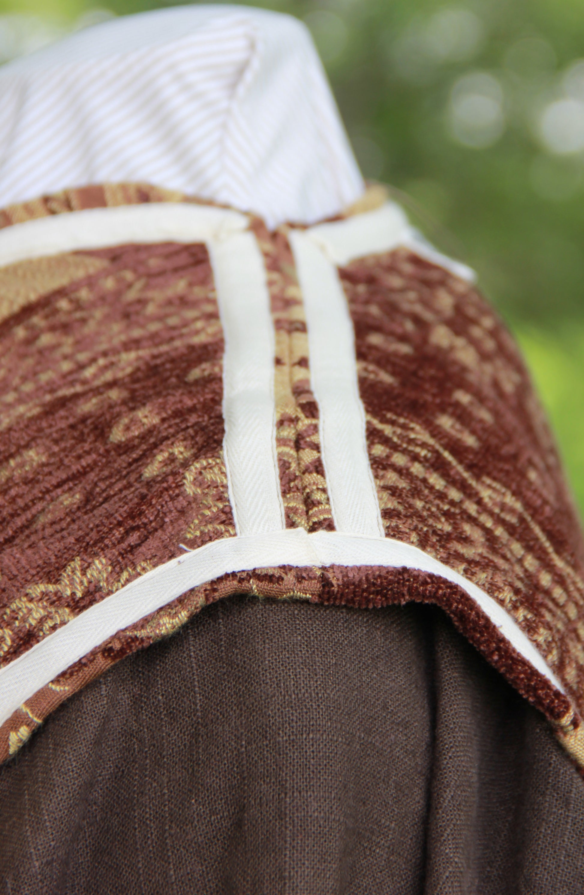 A close up of a brown patterned chenille jerkin at the shoulder. It is inside out to display the ivory twill ribbon running along all of the seams, for decor and function.