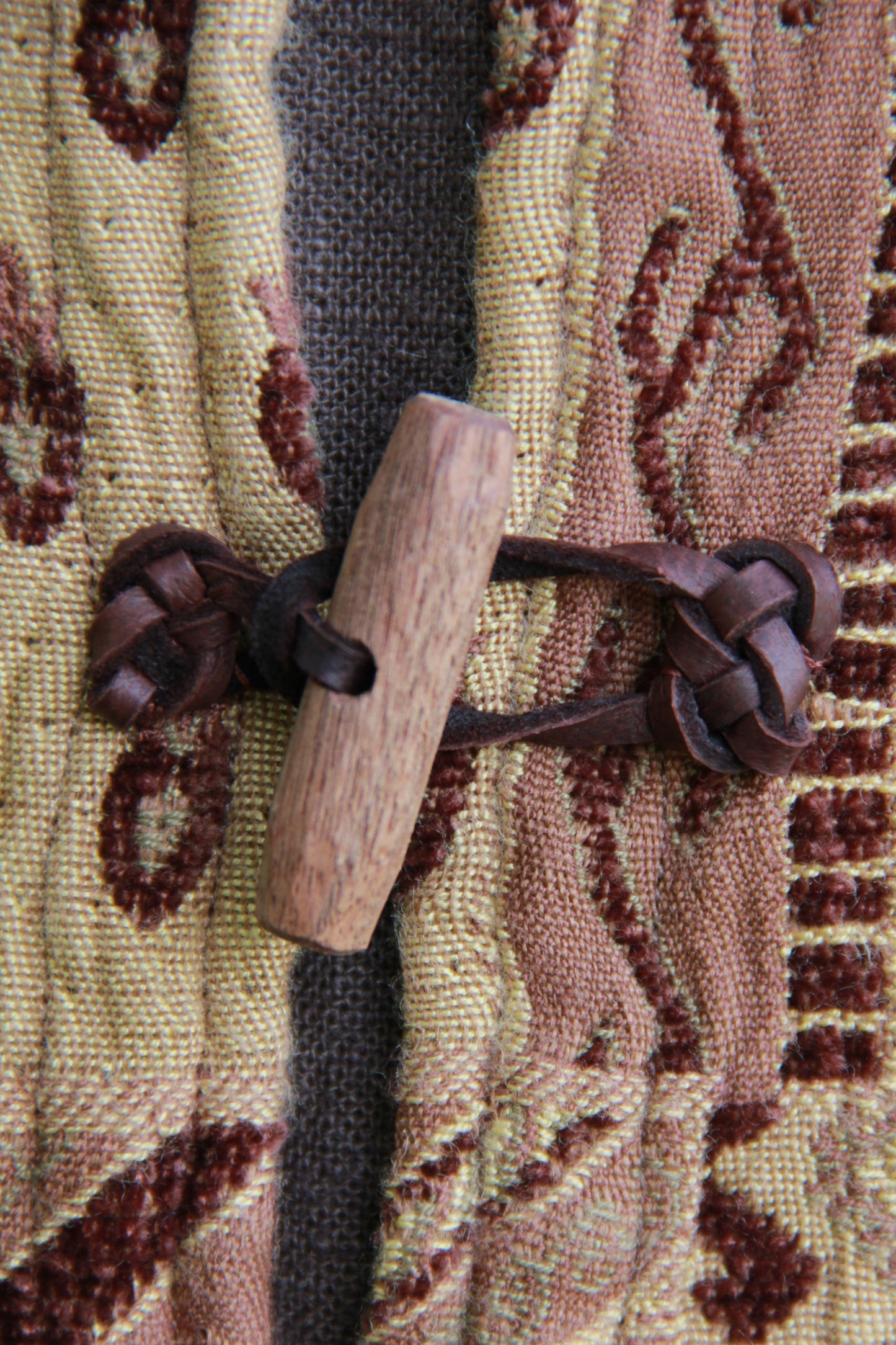 A close up of a hand carved walnut toggle on a tapestry pattered chenille jerkin. The loops securing the toggles are leather with decorative knotwork.