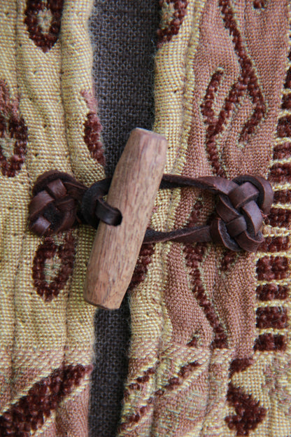 A close up of a hand carved walnut toggle on a tapestry pattered chenille jerkin. The loops securing the toggles are leather with decorative knotwork.