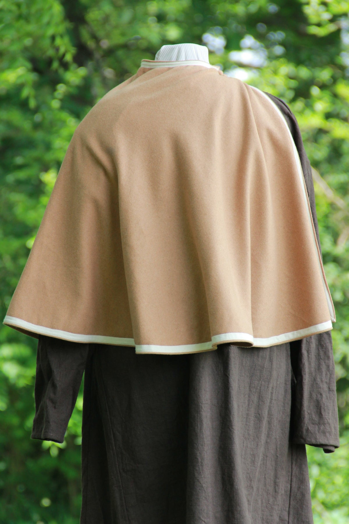A back view of a waist length, semicircle medieval cloak or mantle on a masculine dummy in a woodland setting. It is a camel colored wool and lays open at one shoulder. It is held closed by two buttons made of the same wool and has an ivory ribbon trim around all of the edges. Hand made by west creek craftsman.