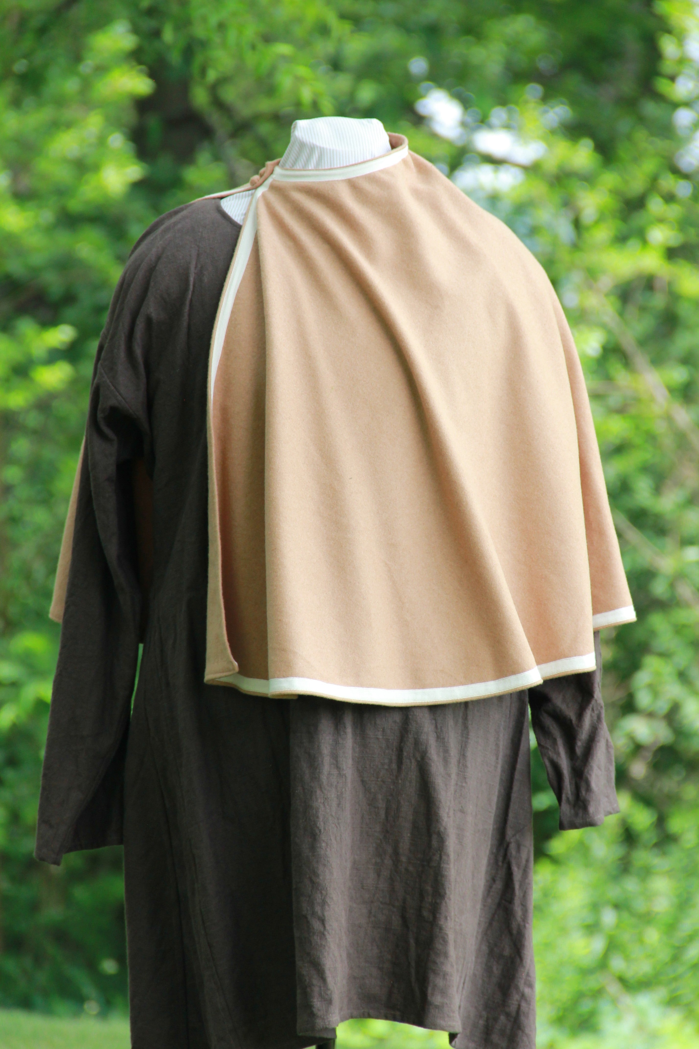 A waist length, semicircle medieval cloak or mantle on a masculine dummy in a woodland setting. It is a camel colored wool and lays open at one shoulder. It is held closed by two buttons made of the same wool and has an ivory ribbon trim around all of the edges. Hand made by west creek craftsman.